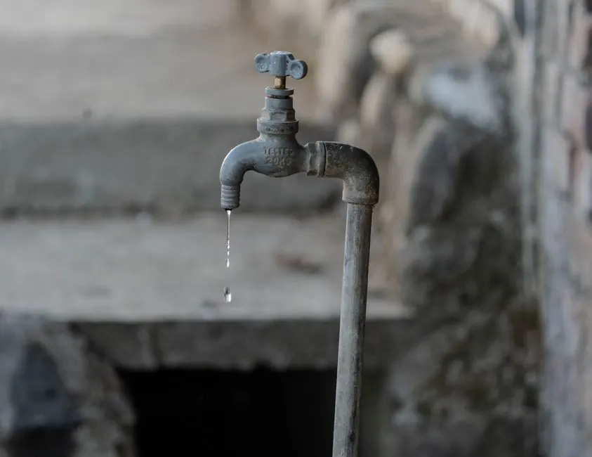 A close-up of a rustic outdoor faucet dripping water against a natural, textured background, symbolizing scarcity.