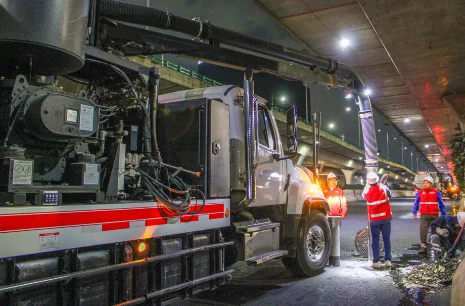 Workers cleaning streets with machinery under an overpass at night.